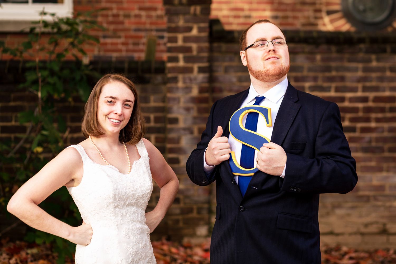 Bride and groom pose humorously with a large letter S prop against a brick wall.