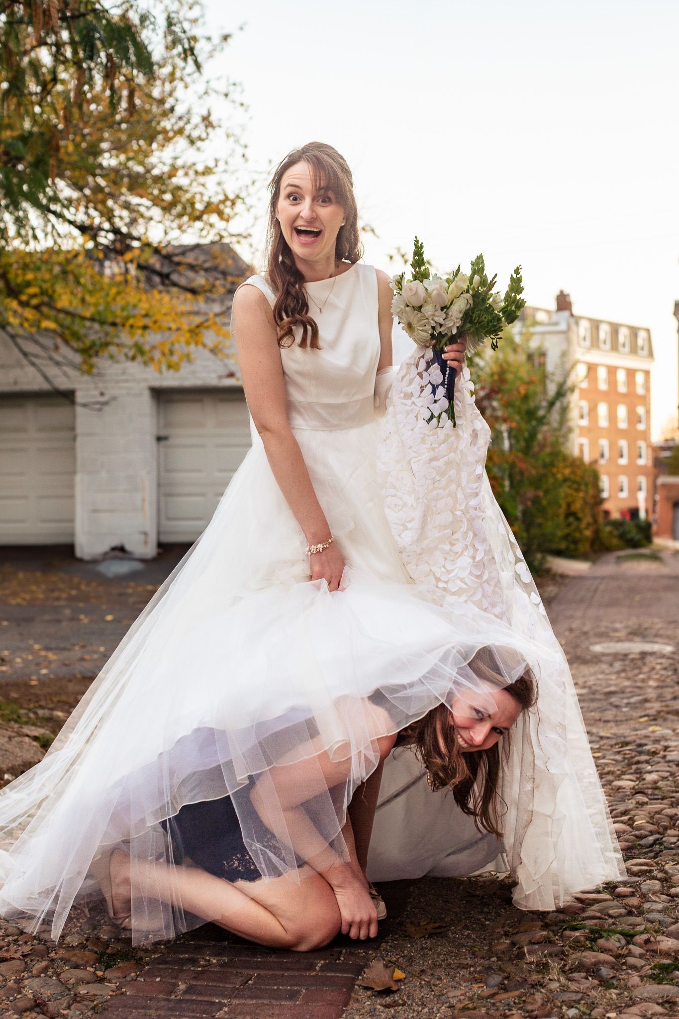 Surprised bride laughing as bridesmaid crawls under her wedding dress holding bouquet.
