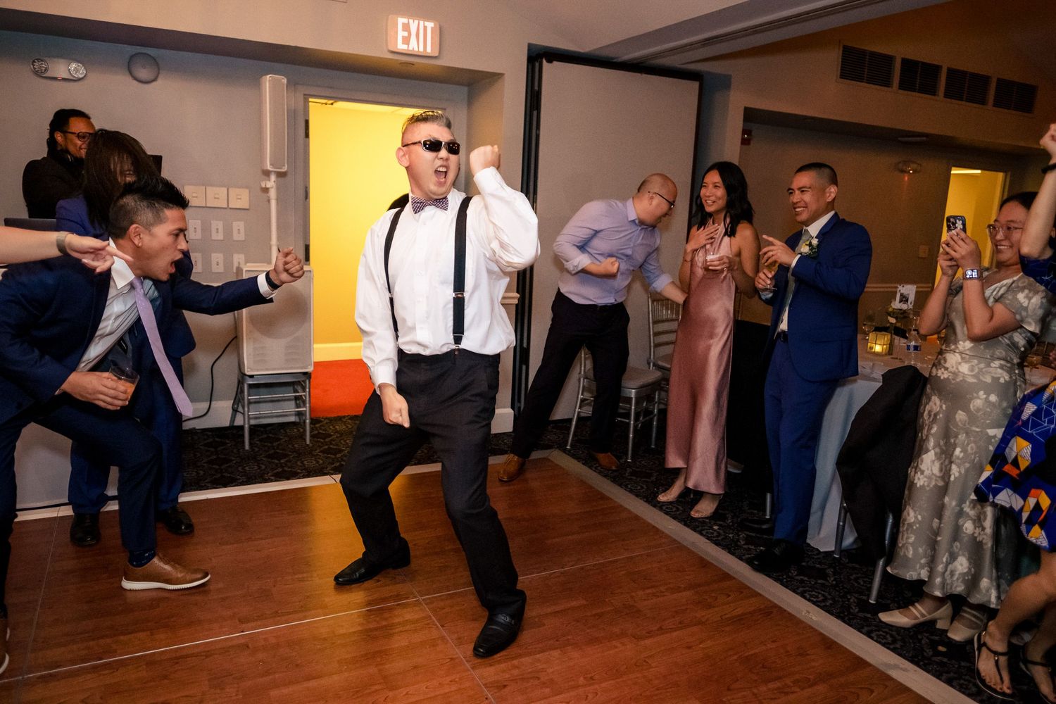 Energetic wedding guest dances in suspenders and sunglasses as guests cheer at the reception.