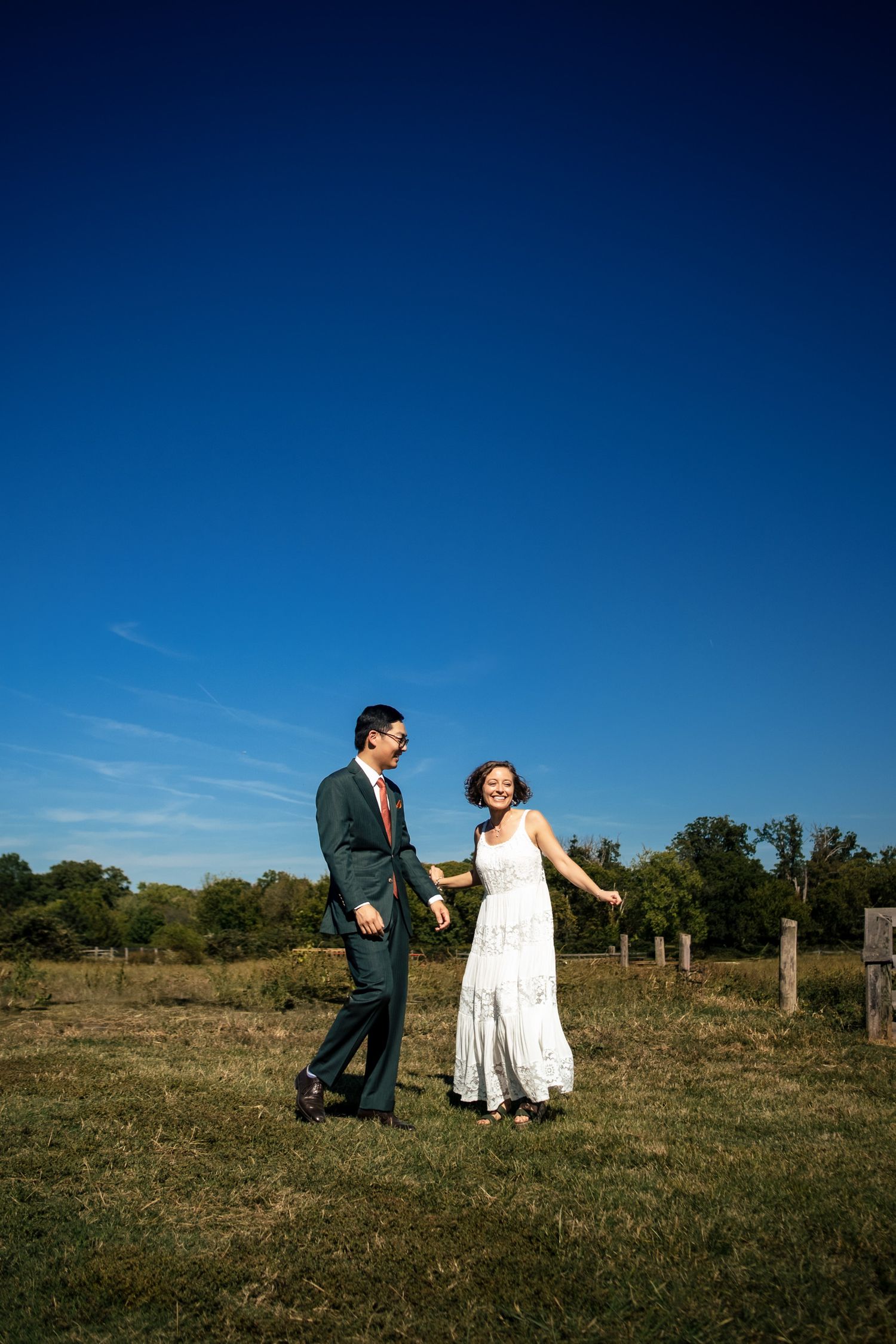 Couple holding hands and laughing in a sunny open field with a vivid blue sky backdrop at their outdoor wedding.