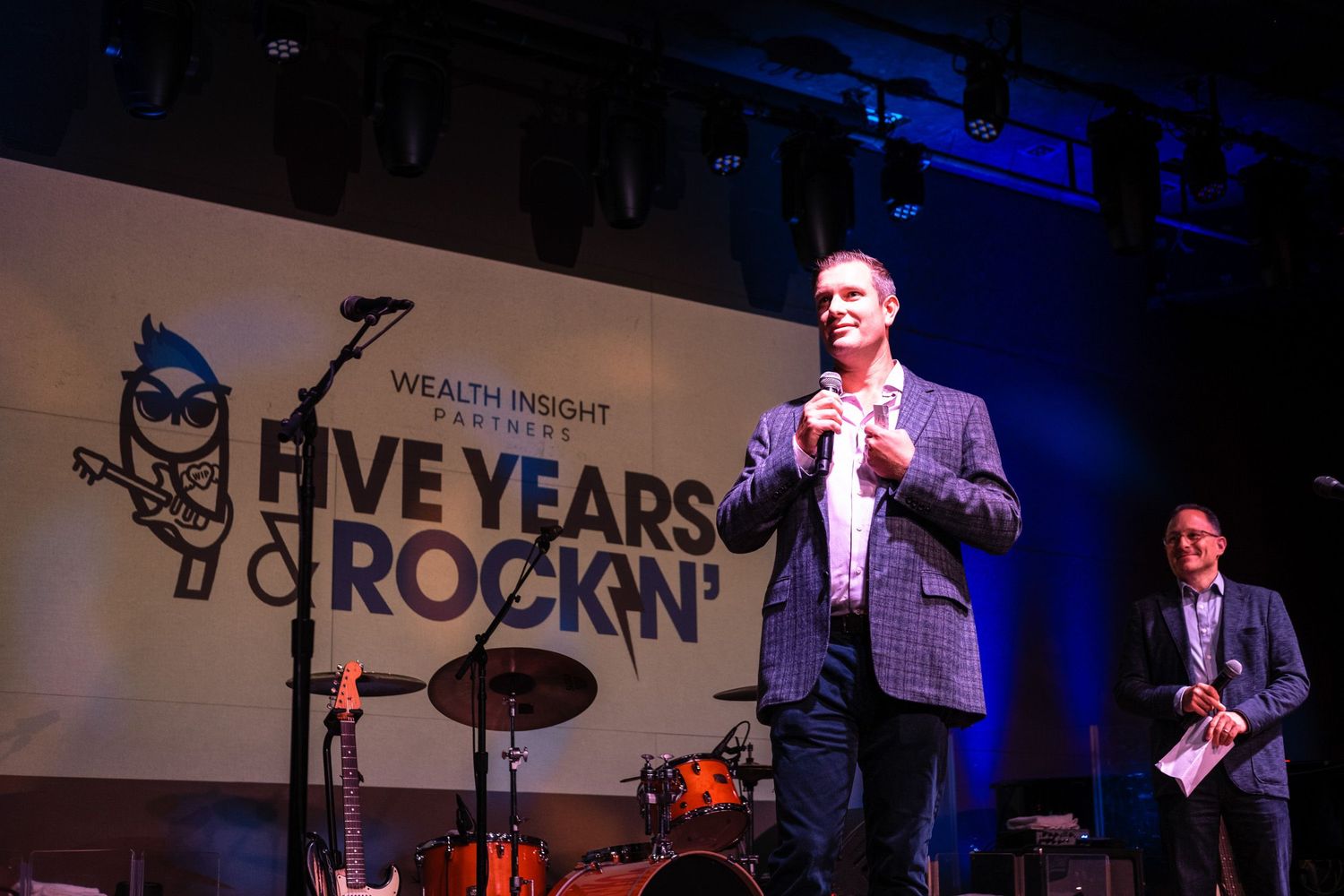 Man speaking on stage at Wealth Insight Partners Five Years Rockin' anniversary event with band setup in background.