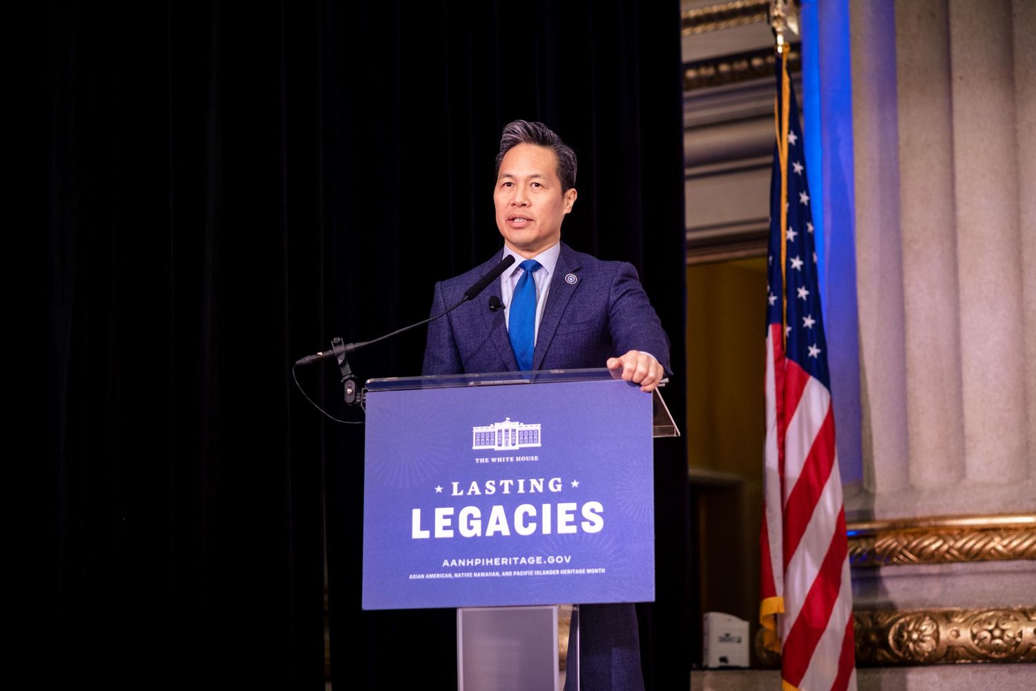Man in blue suit speaks at Lasting Legacies podium with American flag backdrop at formal event.