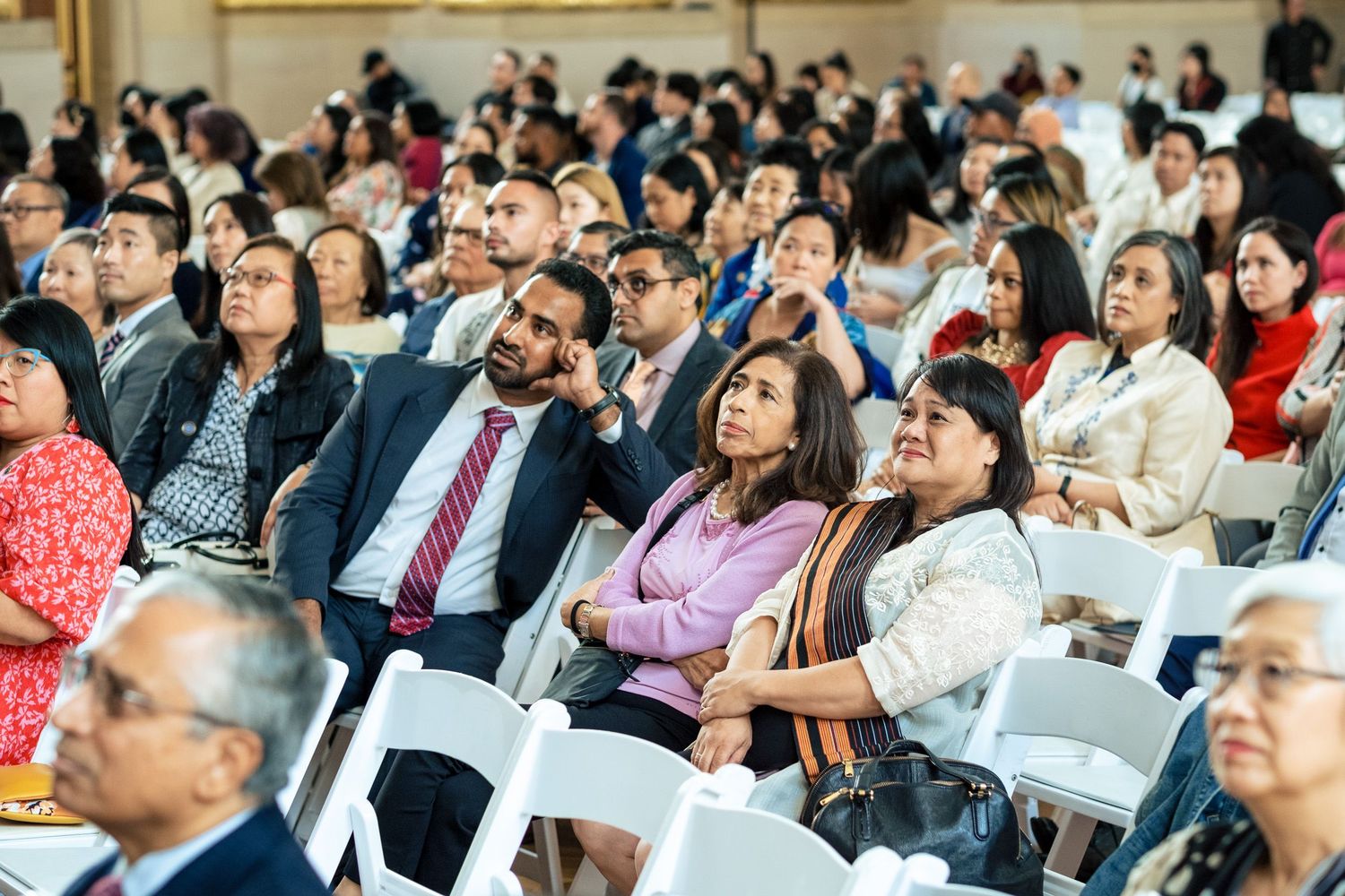 A diverse audience seated in white chairs attends a formal conference or ceremony event.