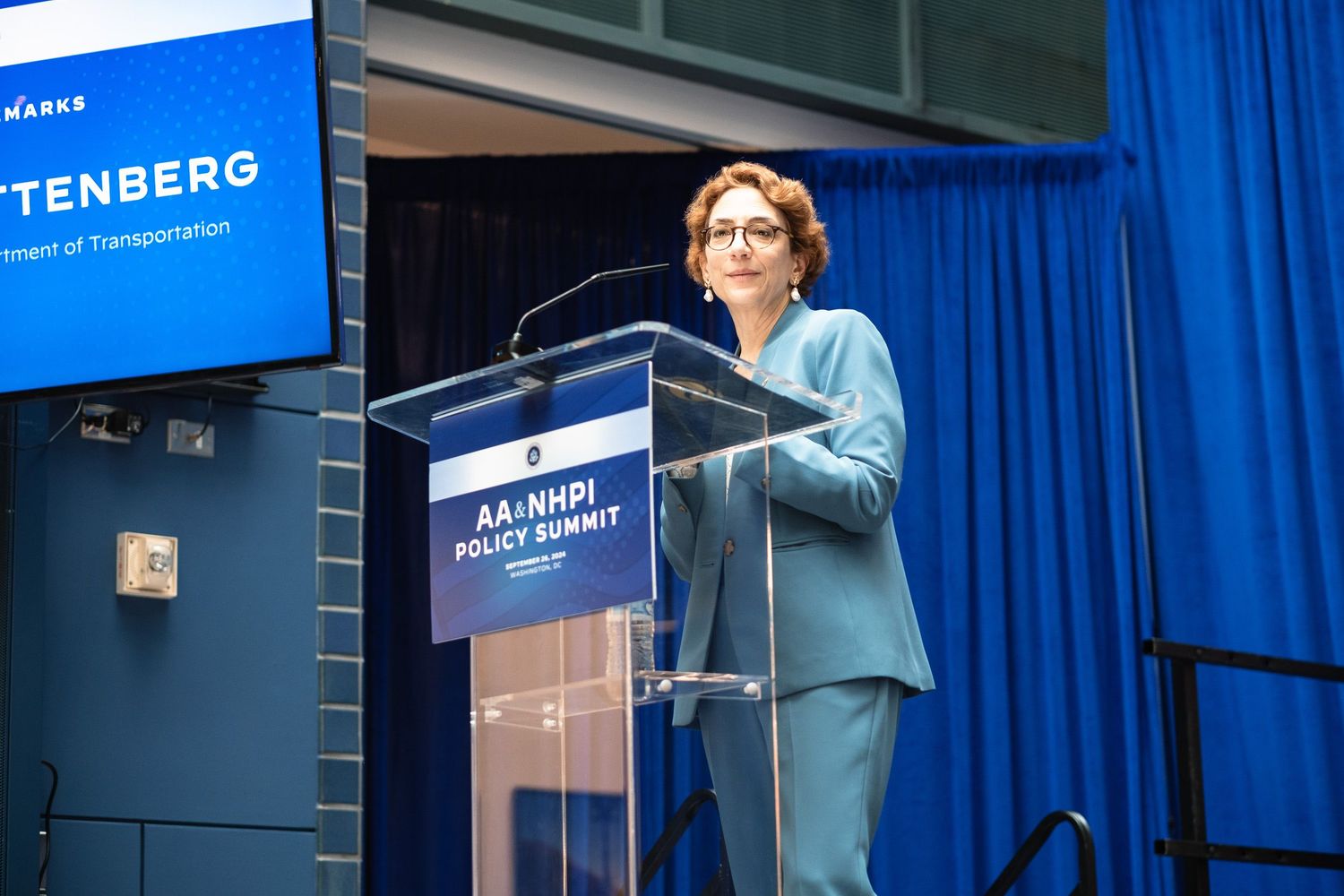 Woman in teal suit speaks at AA-NHPI Policy Summit podium at Department of Transportation event.