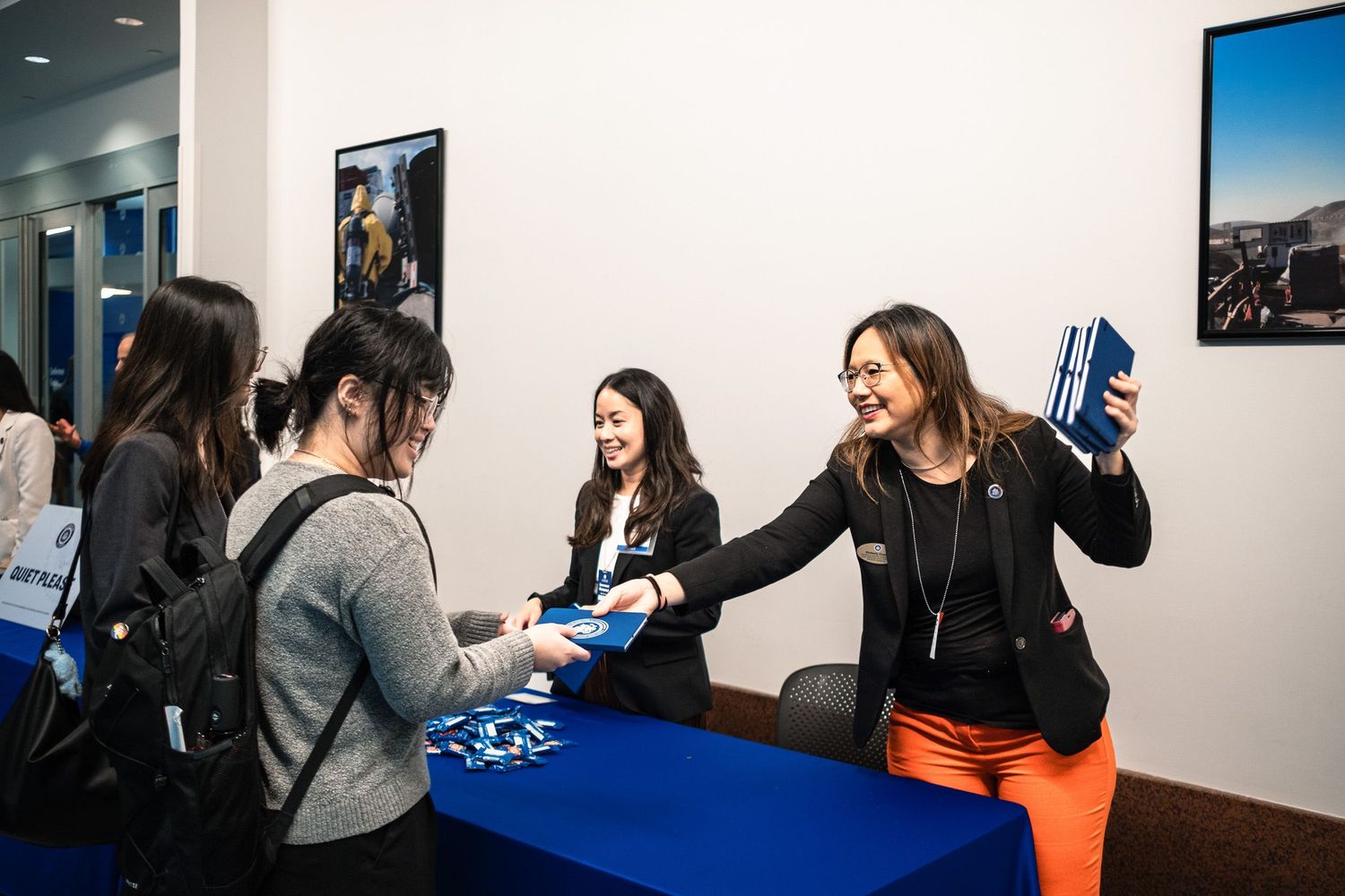 Women at a registration desk exchange blue booklets at a professional conference event.