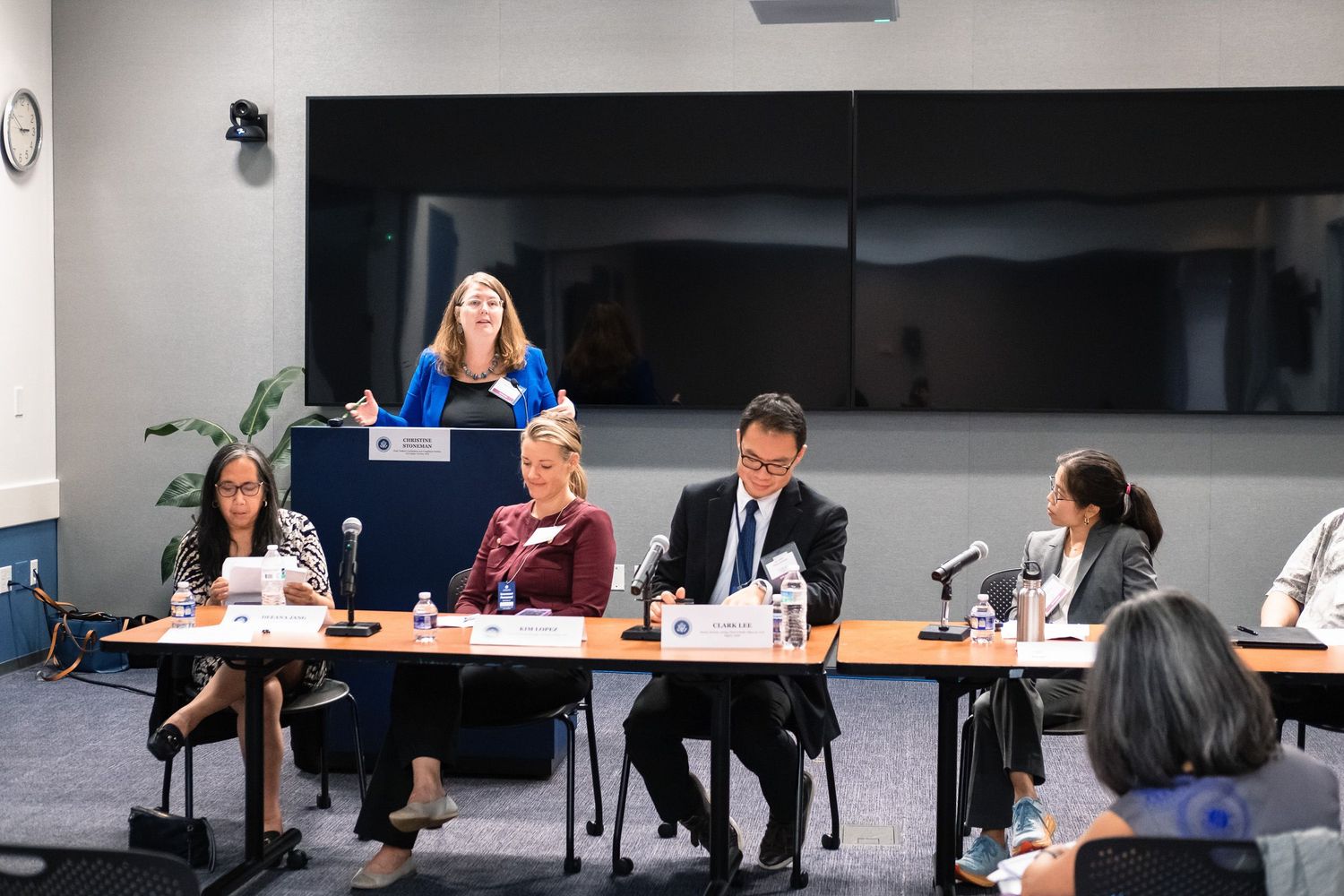 Panel discussion with five professionals at a conference table, one woman standing at podium speaking to seated attendees.