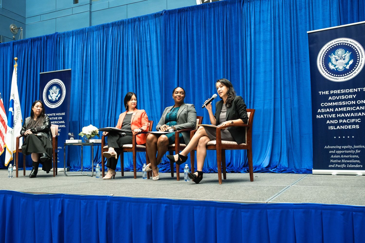 Four women panelists speak on stage at a Presidential Advisory Commission on Asian Americans event with blue drape backdrop.