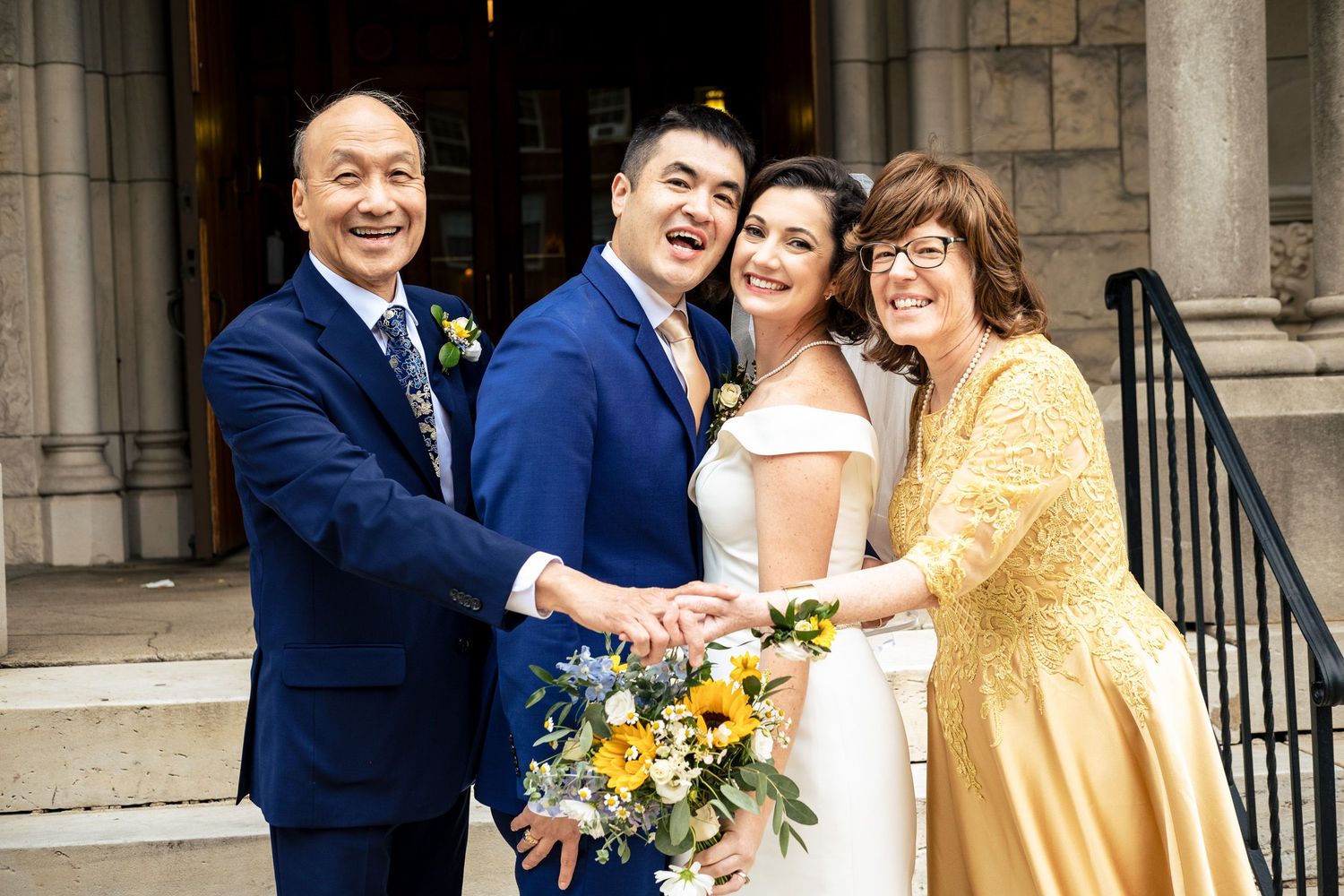Bride and groom celebrating with smiling parents, holding sunflower bouquet, on stone church steps after wedding ceremony.