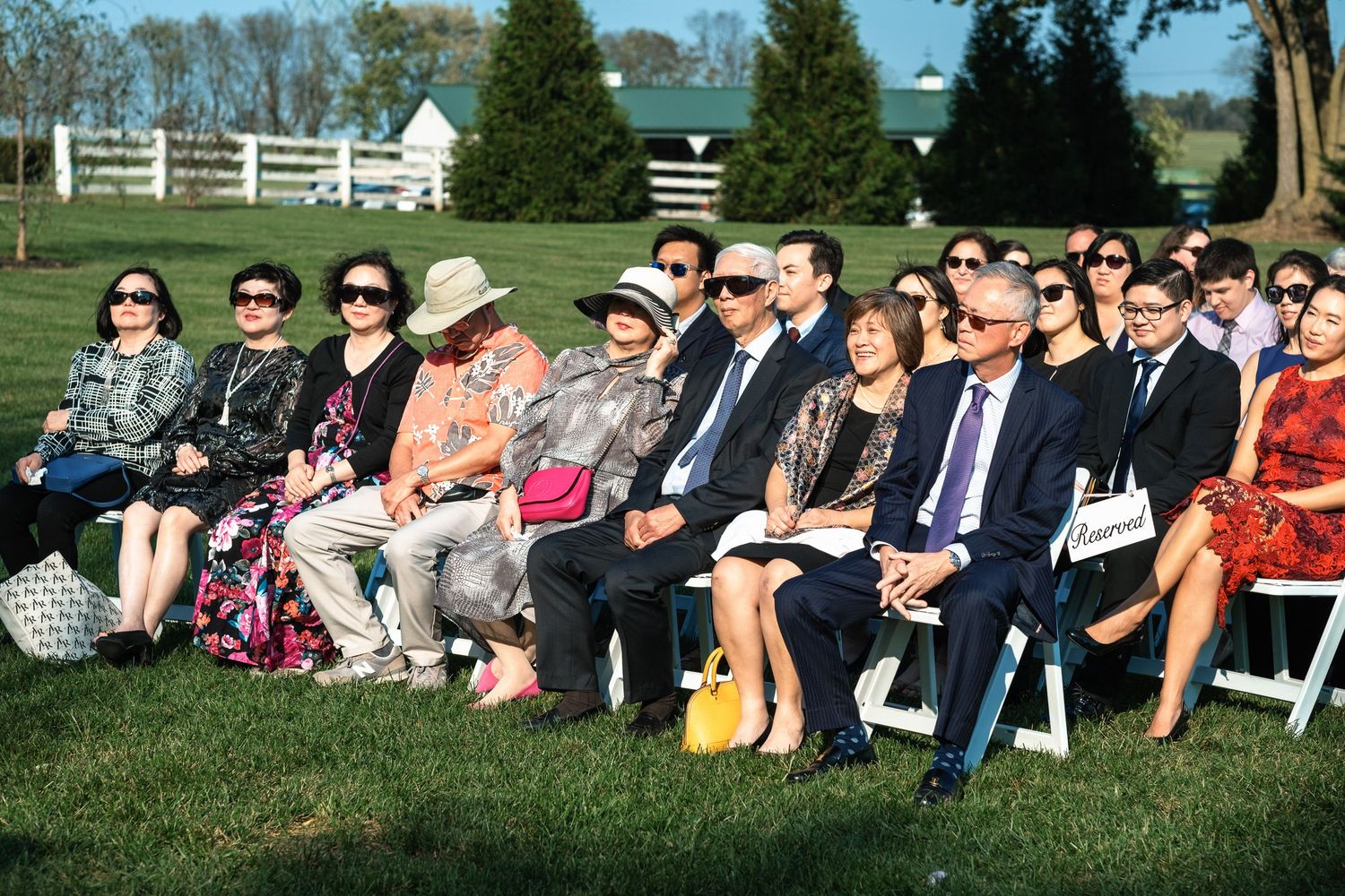 Wedding guests wearing sunglasses seated in white chairs at an outdoor daytime wedding ceremony.
