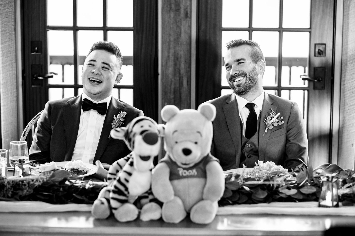 Two grooms laughing joyfully at wedding table with Winnie the Pooh and Tigger stuffed animals.
