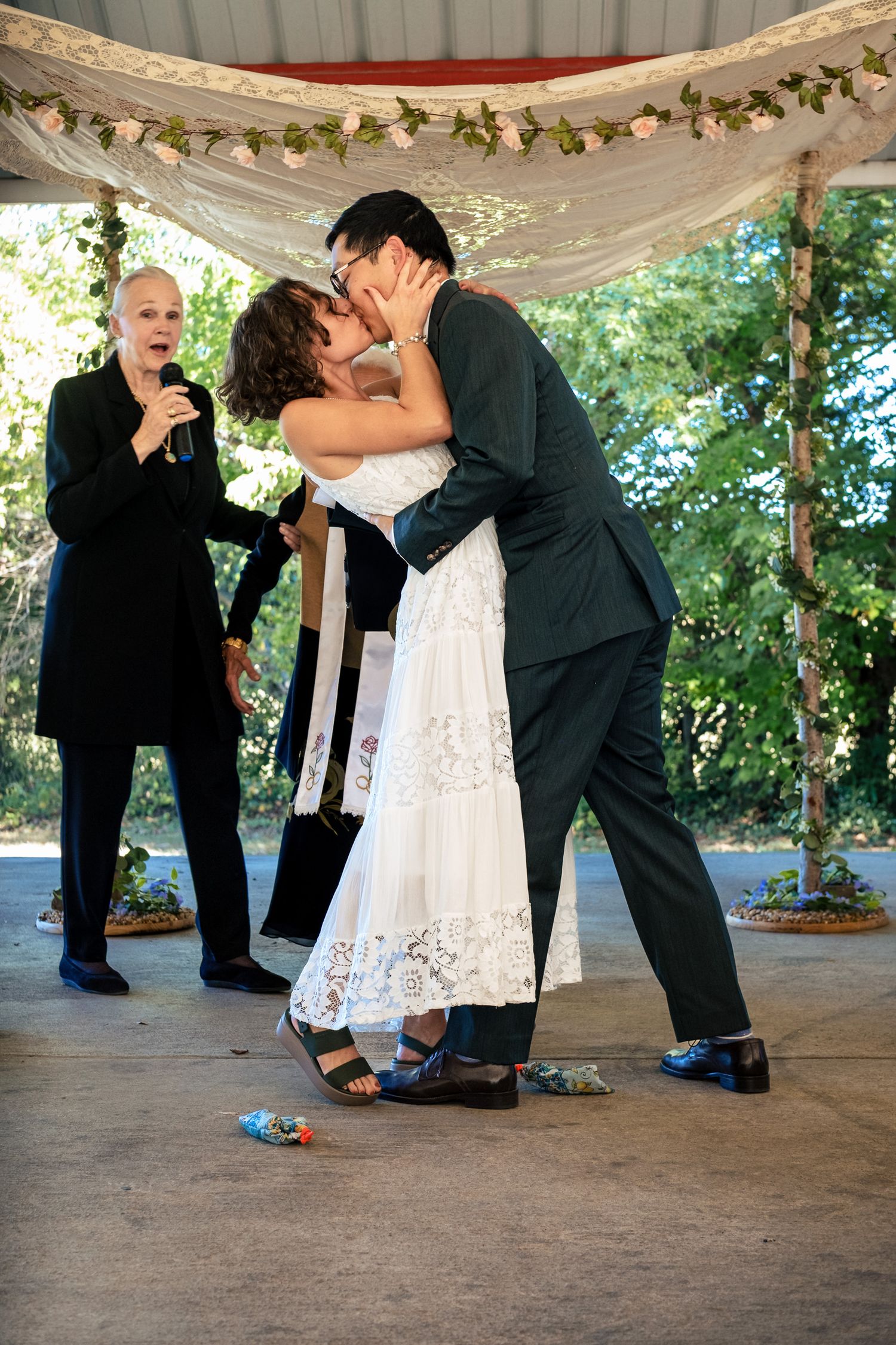 Newlyweds share first kiss at outdoor wedding ceremony while officiant celebrates beside them.
