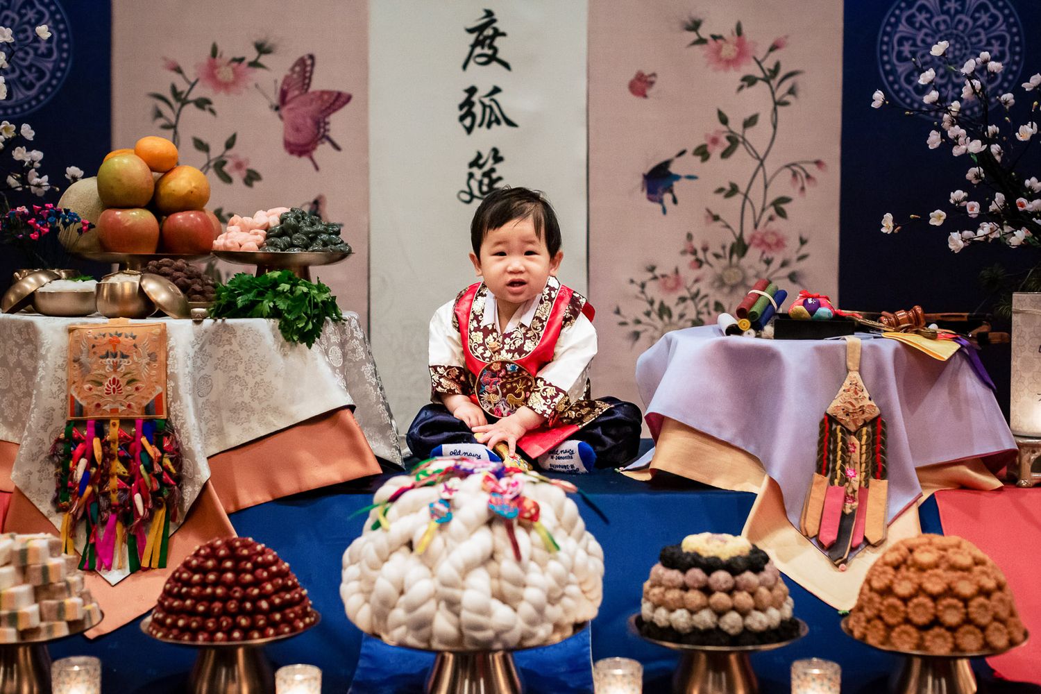 Baby in traditional Korean hanbok sits among ceremonial doljabi food table during first birthday celebration.
