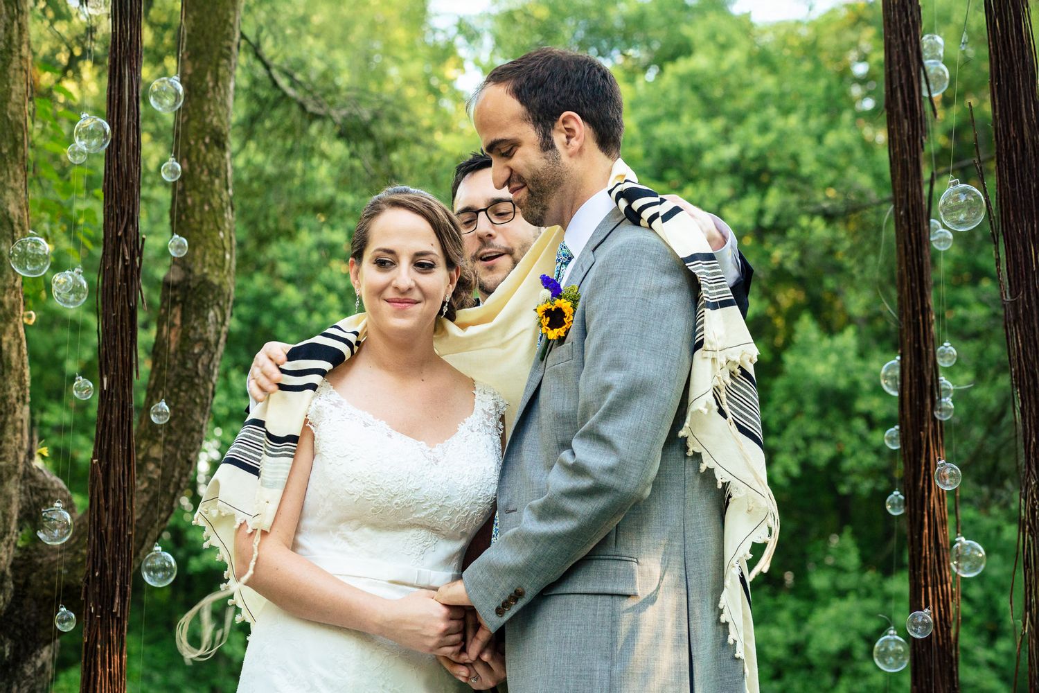 Couple embraces under tallit with officiant during intimate outdoor Jewish wedding ceremony.