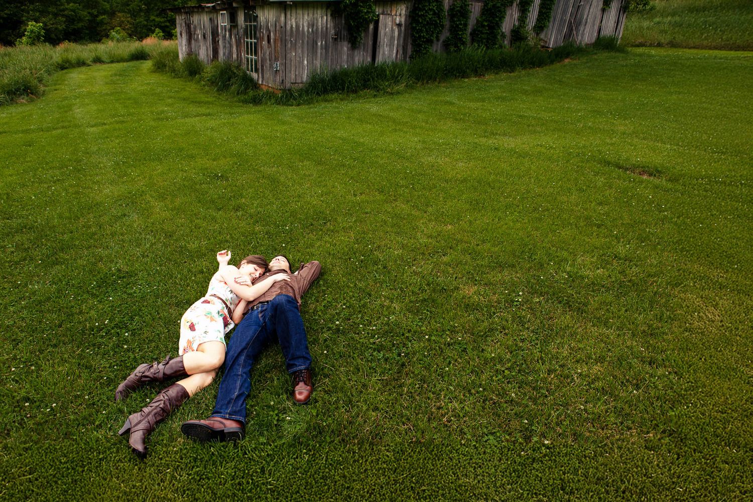 Couple lying together on lush green grass near a rustic wooden fence in countryside.