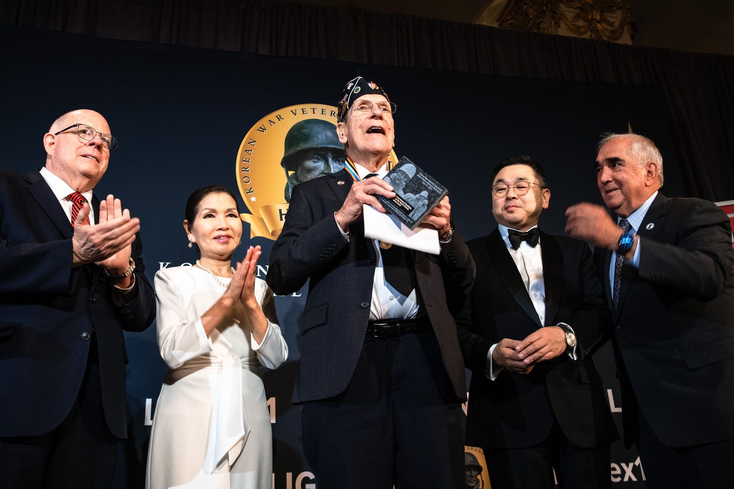 Group of people on stage applauding at a formal awards ceremony with a gold medallion backdrop.