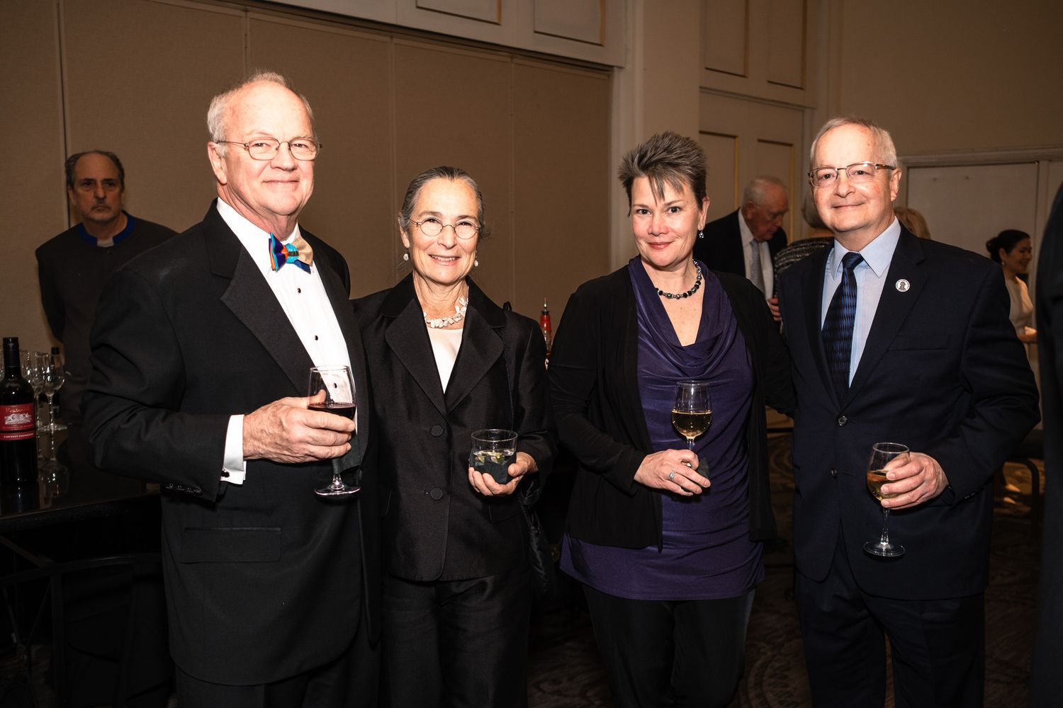 Four formally dressed adults holding wine glasses at an elegant indoor event or gala celebration.