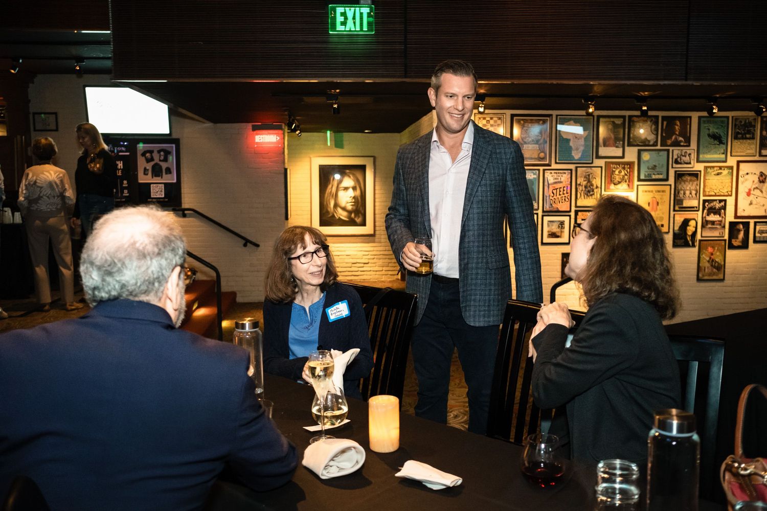 Tall man in plaid blazer networking at indoor restaurant event with guests seated at table near framed artwork.