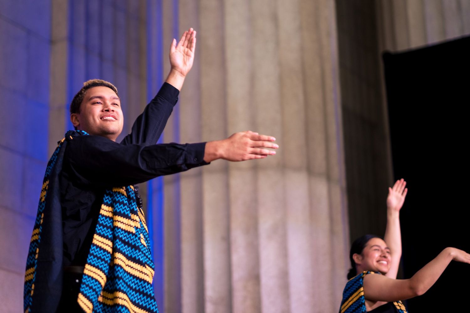 Two performers in colorful African attire dance gracefully on an outdoor stage with columns in the background.