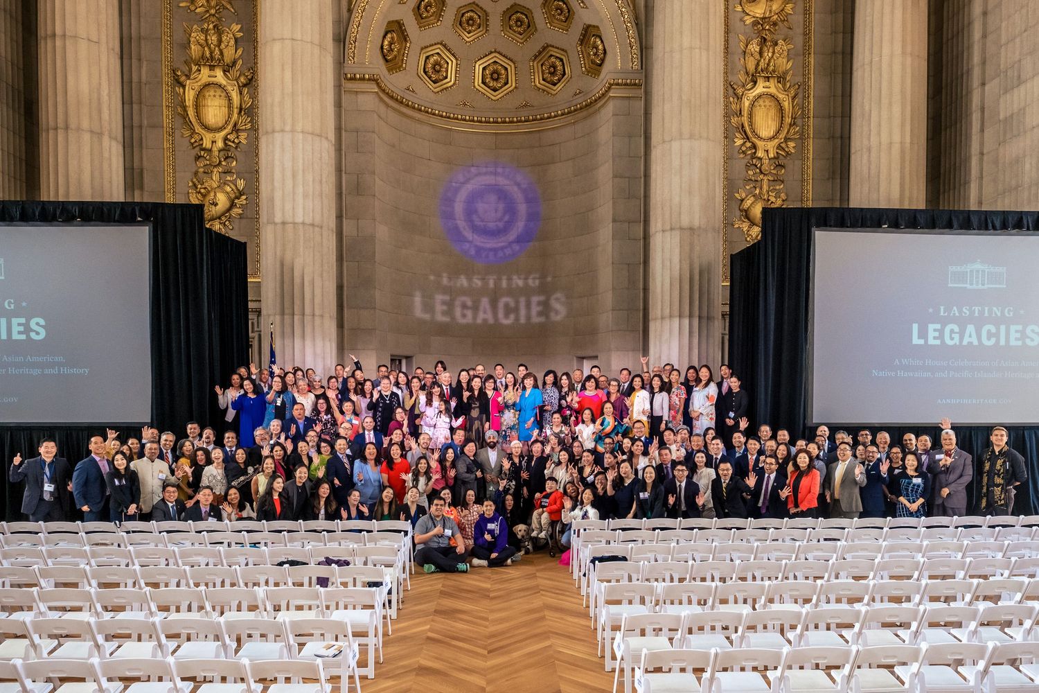 Large group photo at Lasting Legacies event inside grand hall with ornate columns and rows of white chairs.
