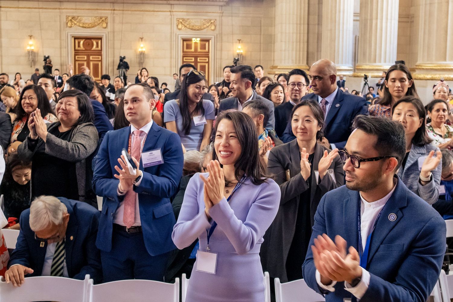 Diverse professionals applauding and smiling at a formal event in an elegant hall, wearing business attire and name badges.
