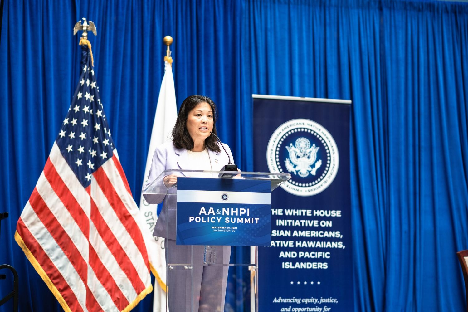 Woman speaking at podium during AANHPI Policy Summit with White House Initiative banner and American flag backdrop.