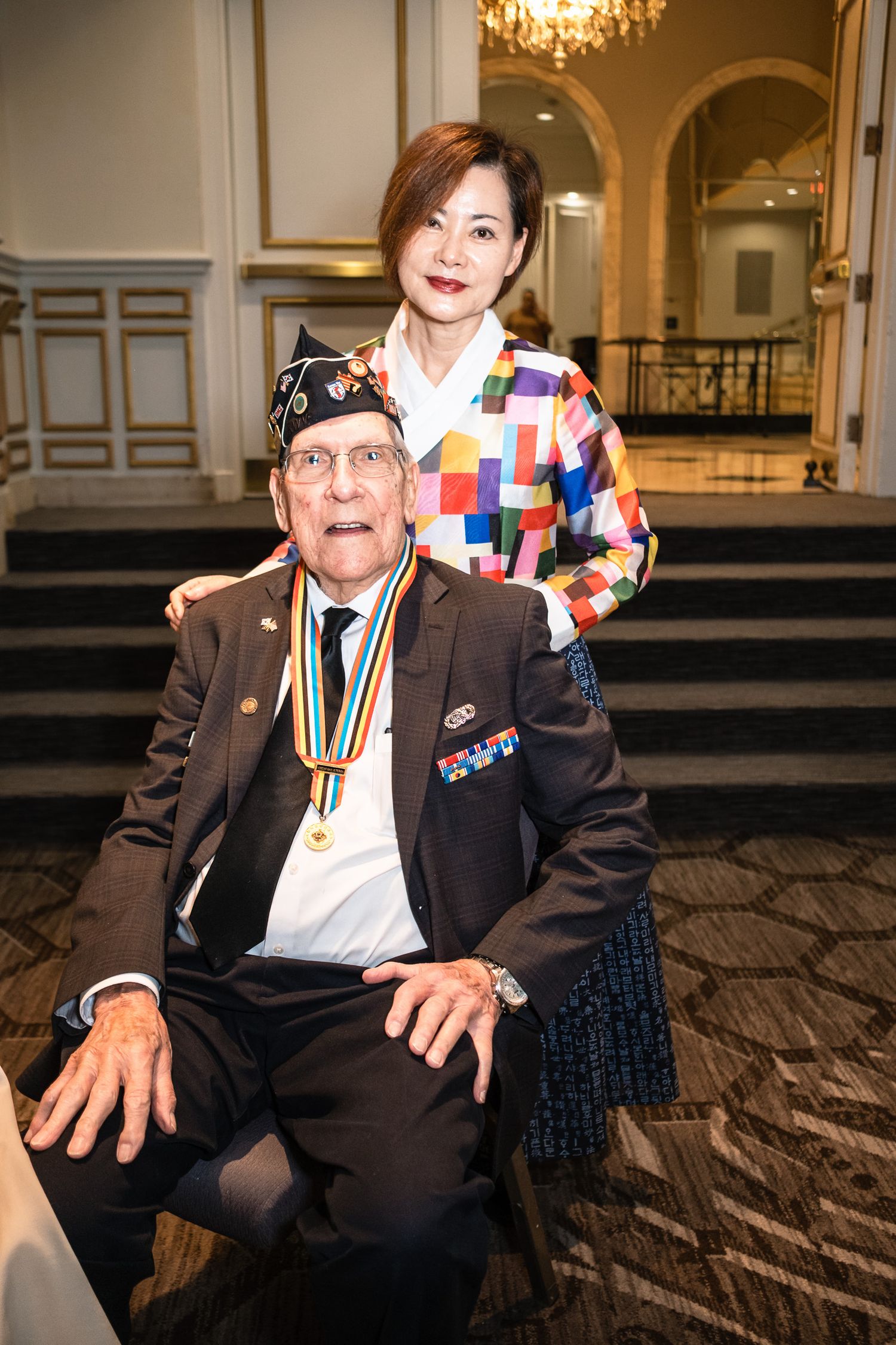 Elderly veteran wearing medals and military cap poses with a woman in colorful jacket in an elegant ballroom.