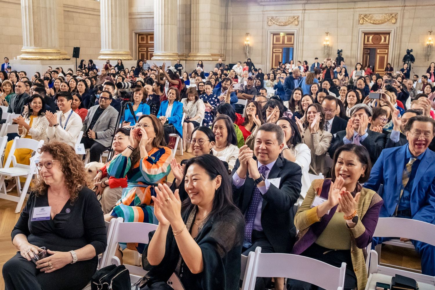 A diverse crowd of people clapping and smiling at an indoor event in an elegant hall.