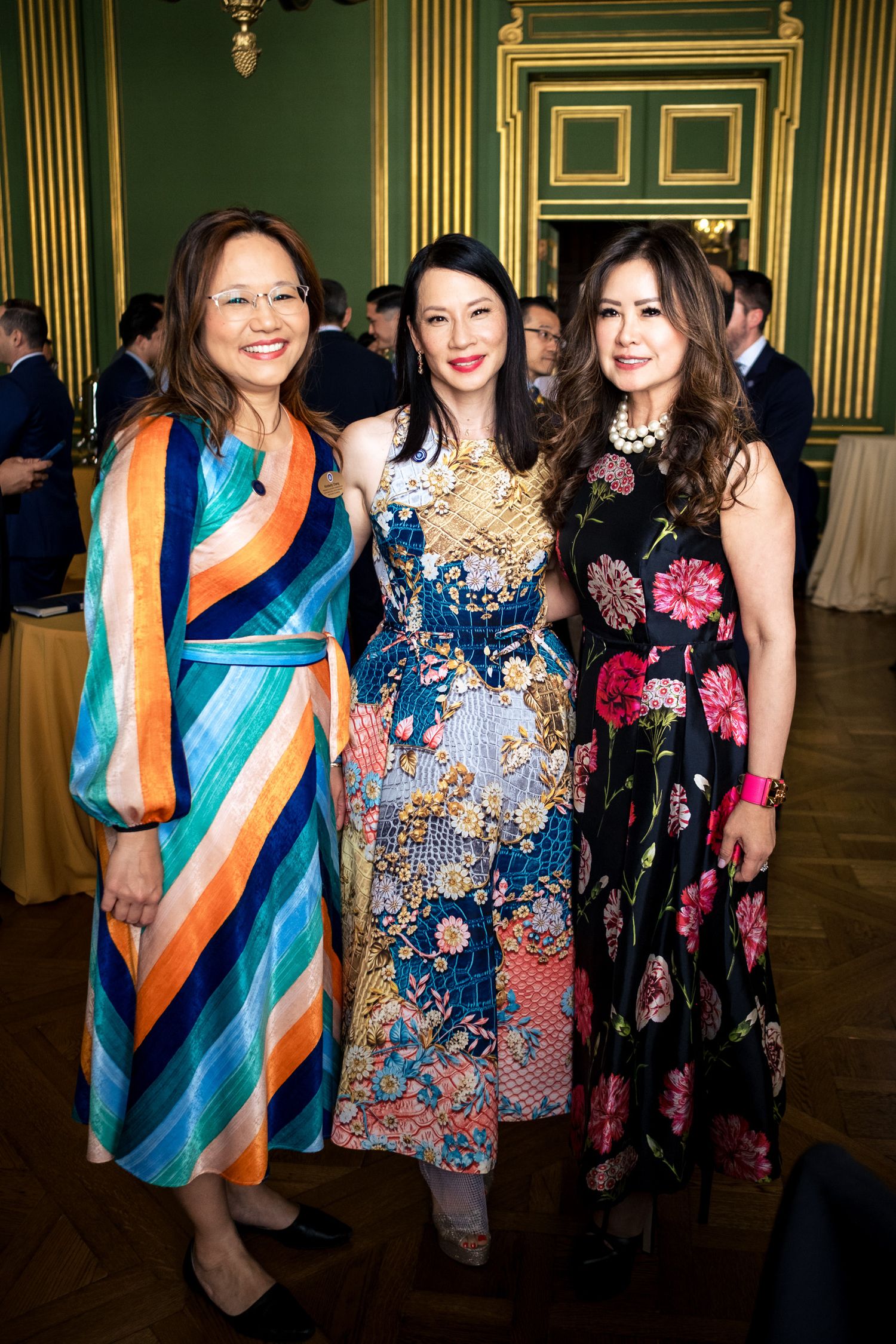 Three elegantly dressed women pose together at a formal event, wearing colorful floral and striped gowns.