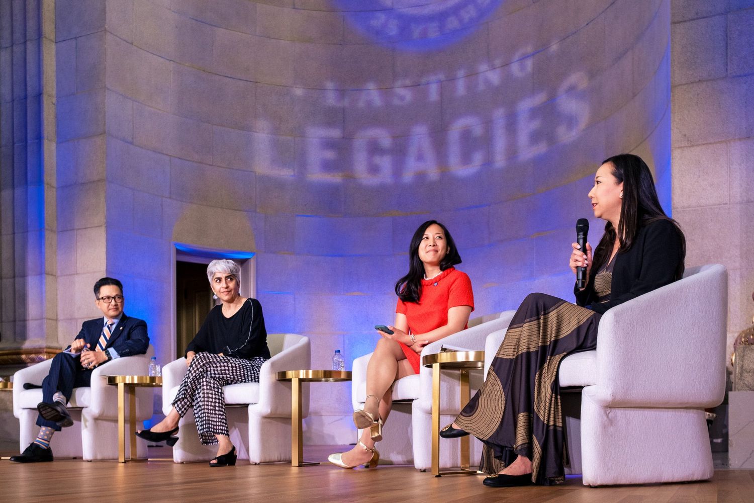 Four panelists speak on stage at a Lasting Legacies event, seated in white chairs under blue lighting.
