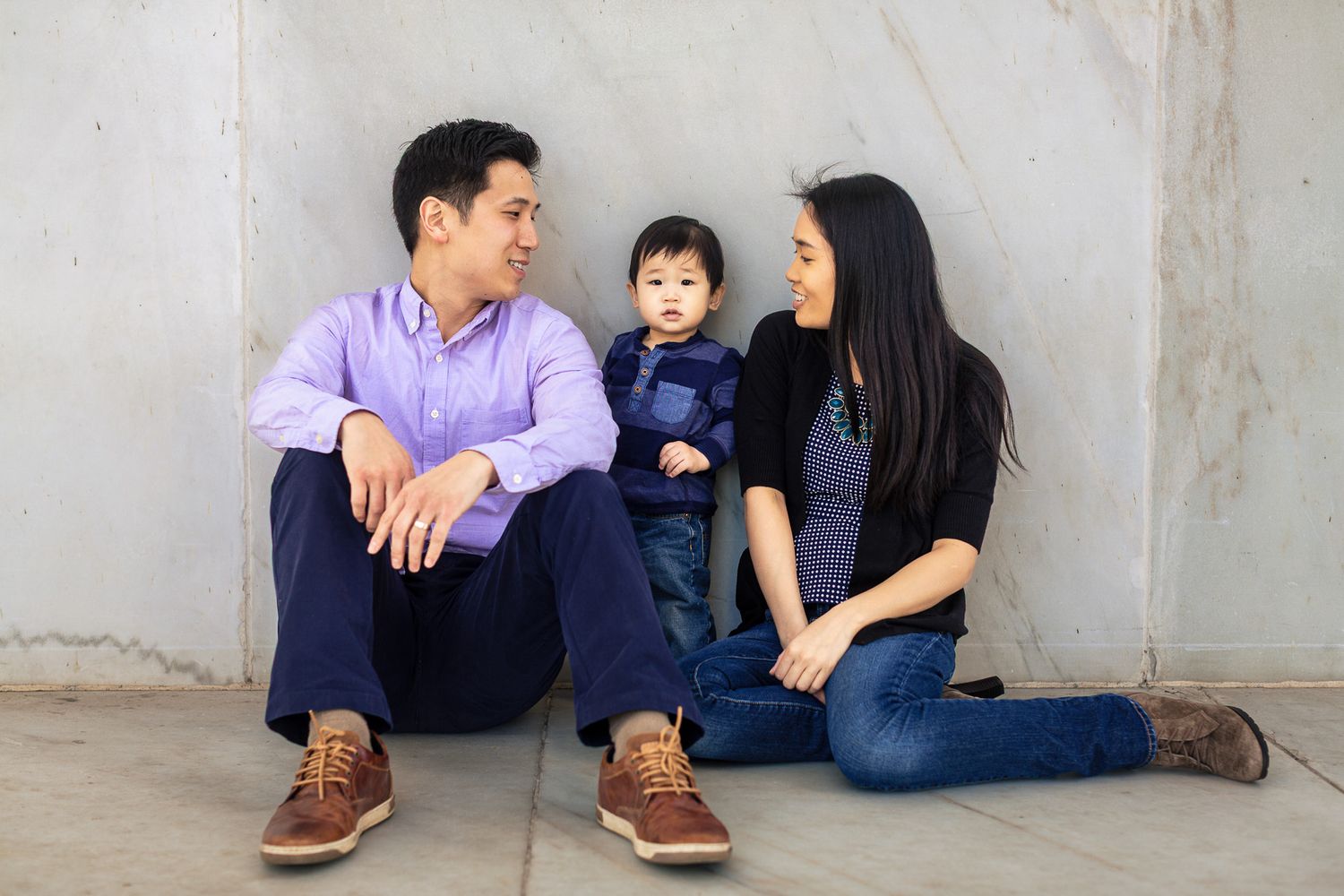 Young Asian family of three sitting casually together against a concrete wall smiling warmly.