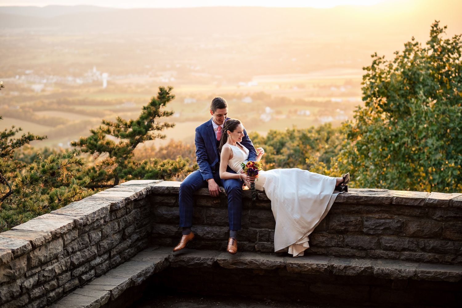 Bride and groom sitting on a stone wall at sunset with a scenic valley view.