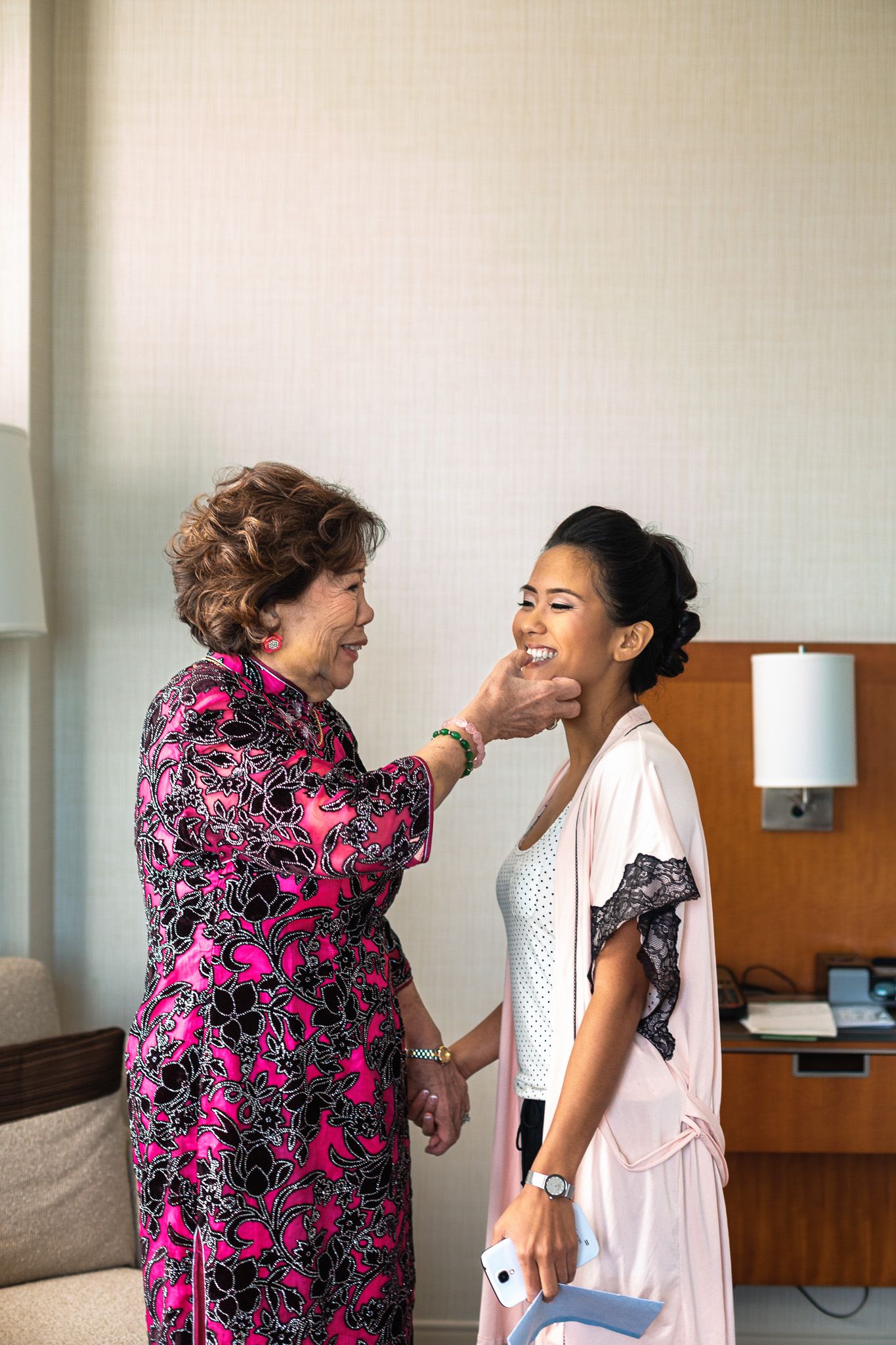 Older woman in pink floral dress tenderly touches the face of a smiling bride-to-be.