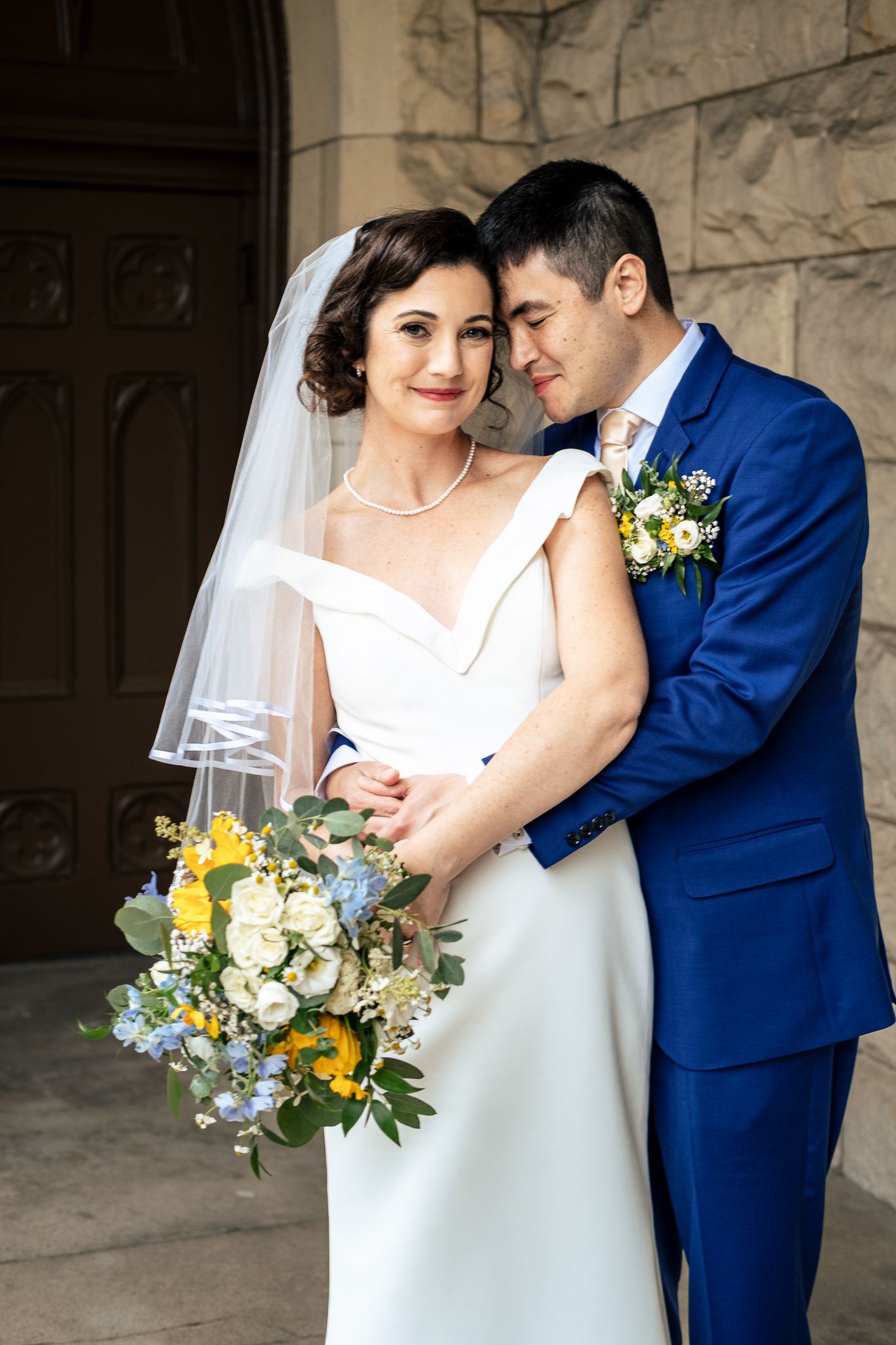 Groom in blue suit embraces bride holding yellow and white bouquet outside a stone church entrance.