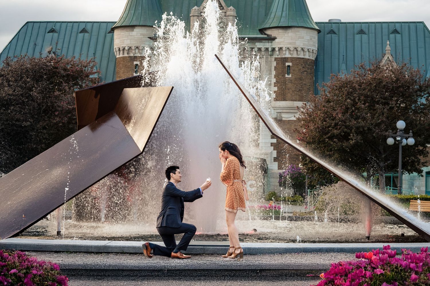 Man proposing on one knee to woman near a fountain and castle-like building outdoors.