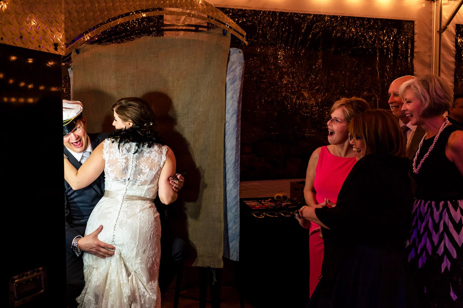 Bride in lace dress hugging groom at wedding photo booth while smiling women watch nearby.