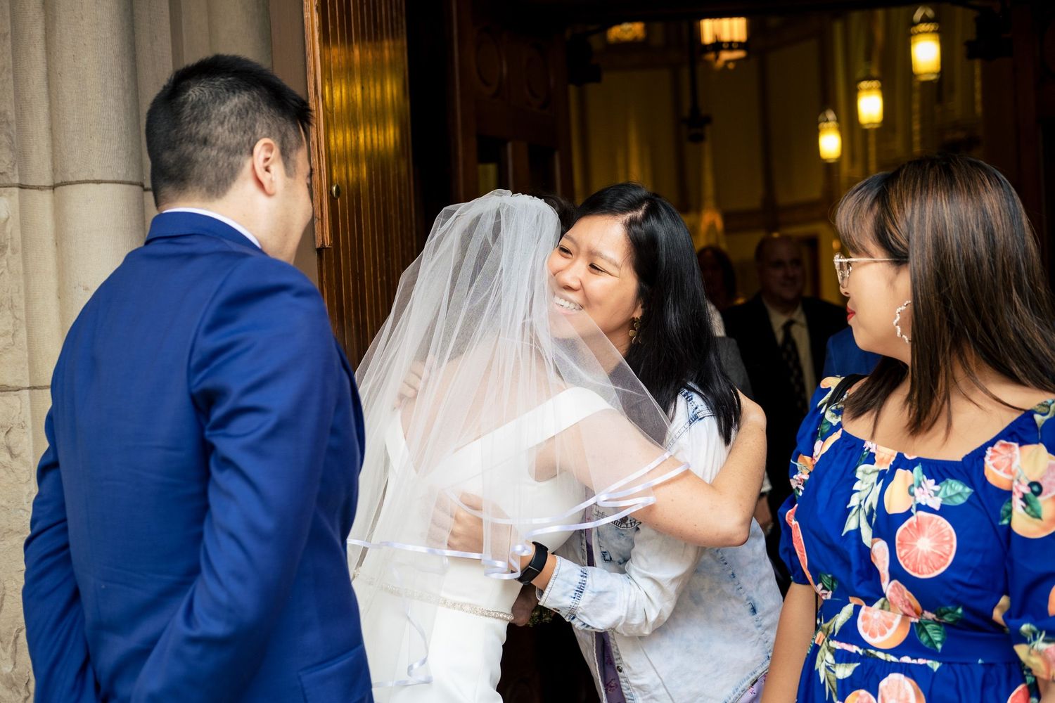 Smiling wedding guest embracing bride in white gown and veil while another guest in floral dress looks on warmly.