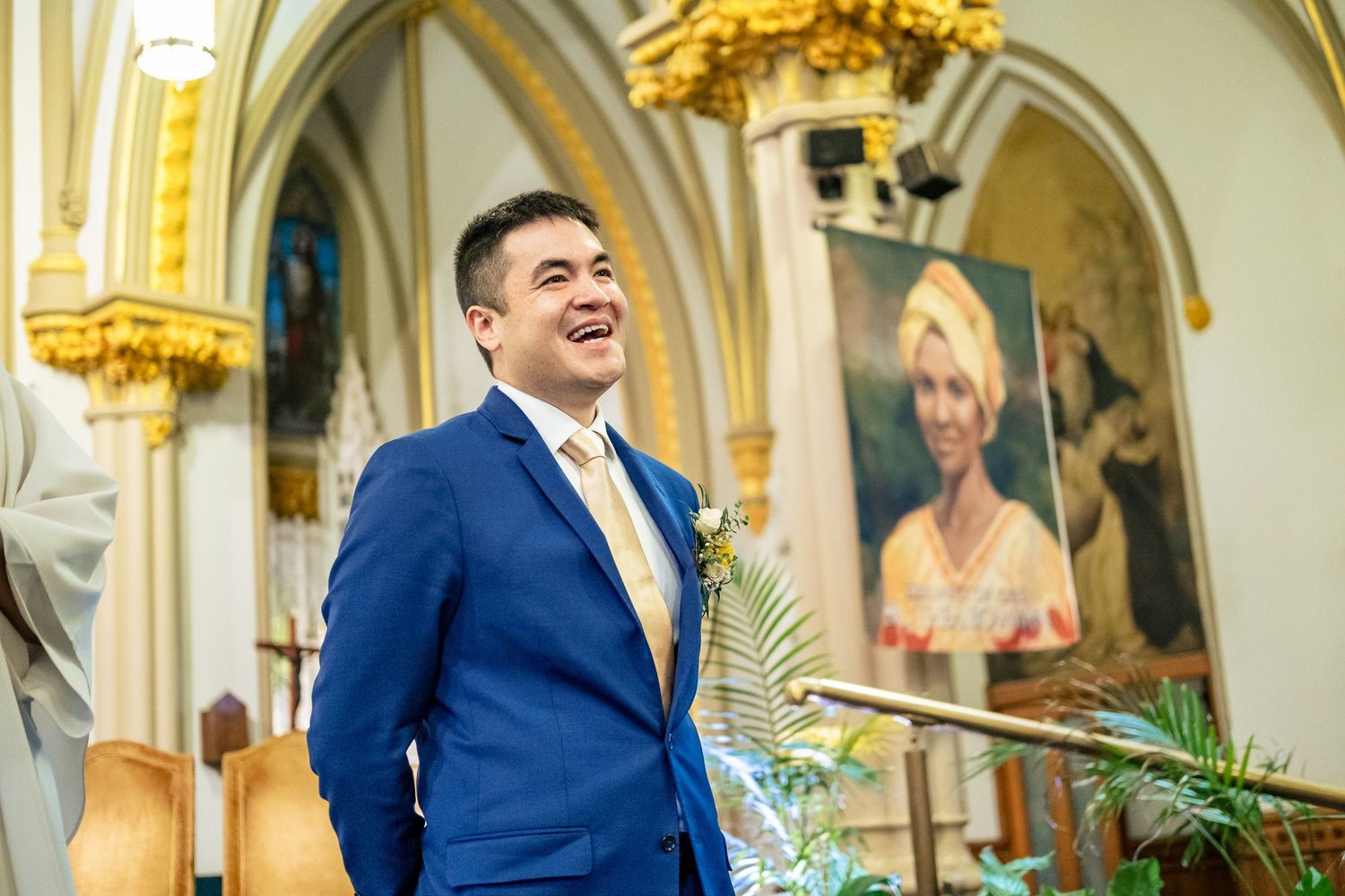 Smiling groom in blue suit stands at church altar with floral boutonniere during wedding ceremony.