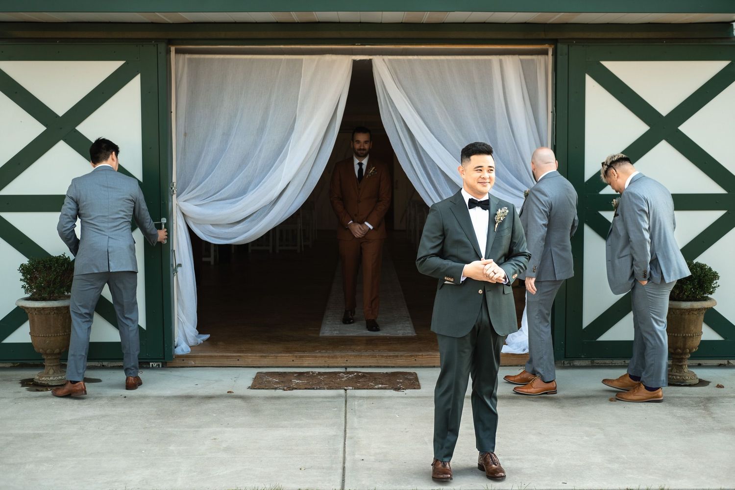 Groom in black tuxedo stands in front of barn as groomsmen emerge through draped doorway.