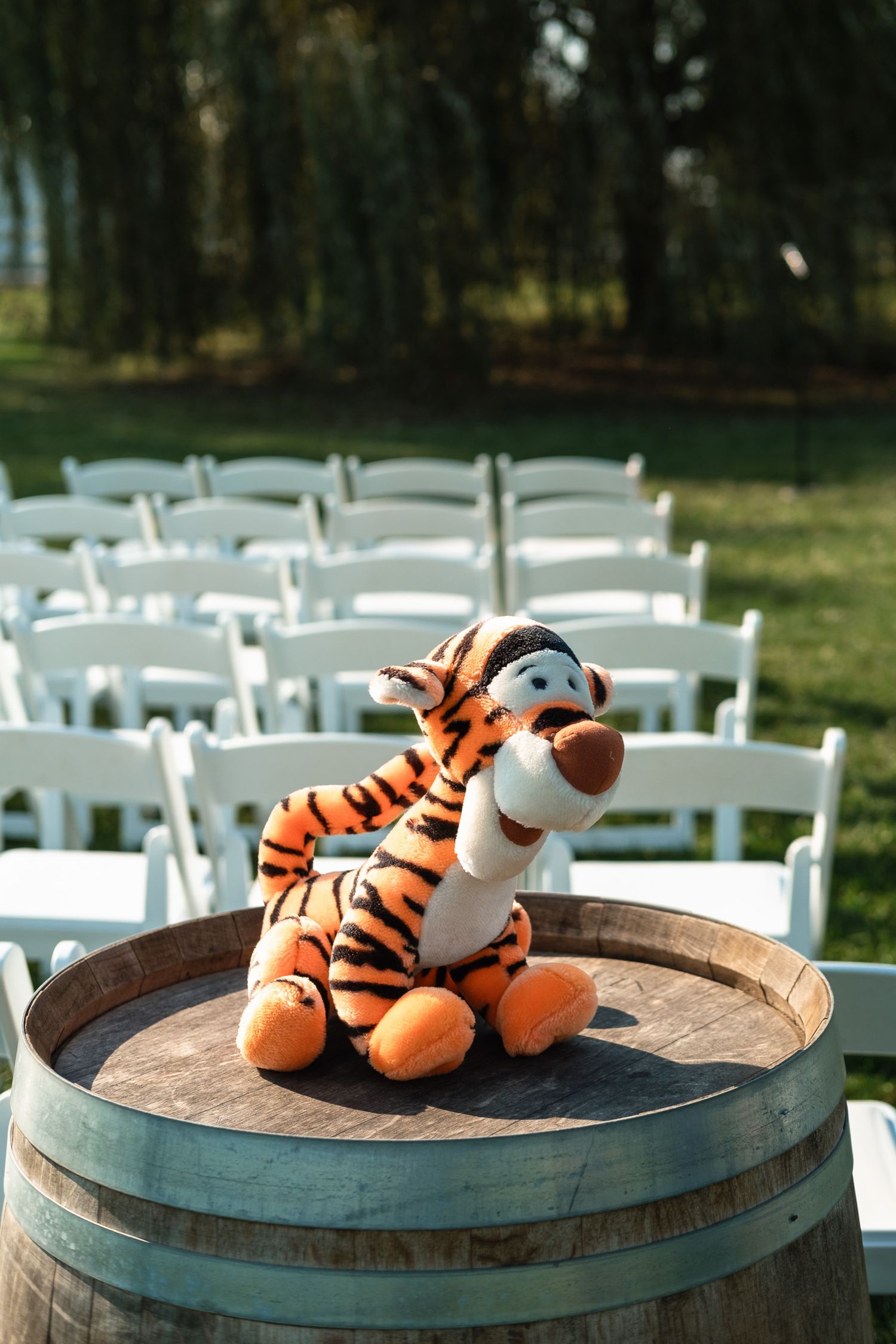 Tigger stuffed animal sits on a wooden barrel beside white chairs at an outdoor wedding ceremony.