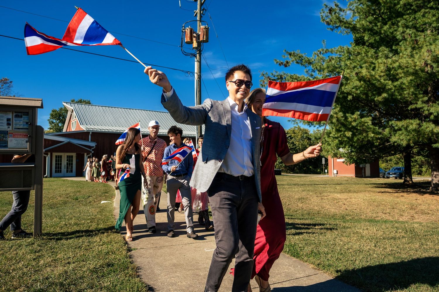 A man in a suit leads a festive procession waving a Thai flag on a sunny outdoor wedding day.