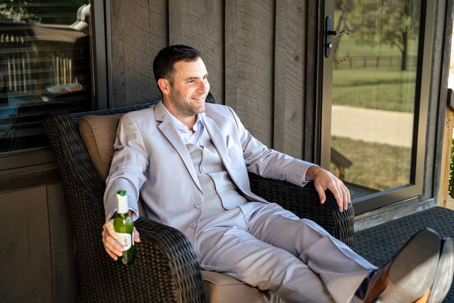 Groom in light gray suit relaxing in wicker chair holding a beer bottle on a porch.