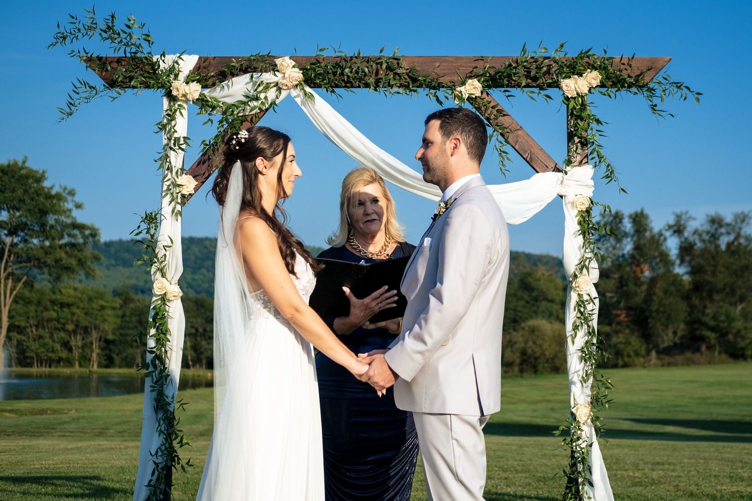 Bride and groom hold hands before an officiant under a floral arch during an outdoor wedding ceremony.