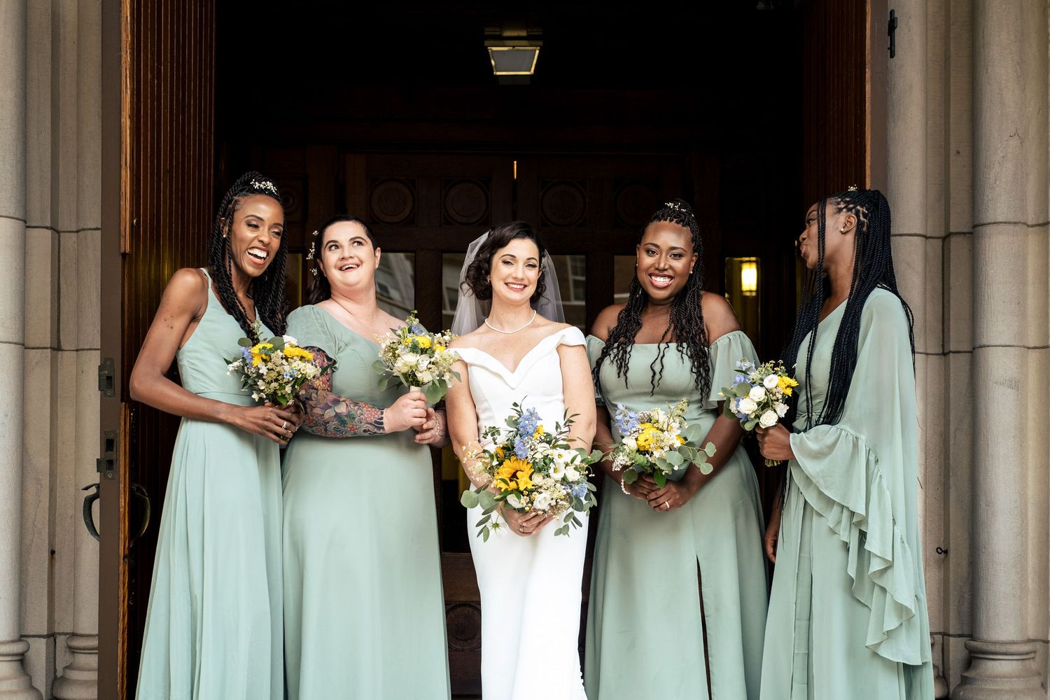 Bride poses with four bridesmaids in sage green dresses holding yellow and white bouquets at church.