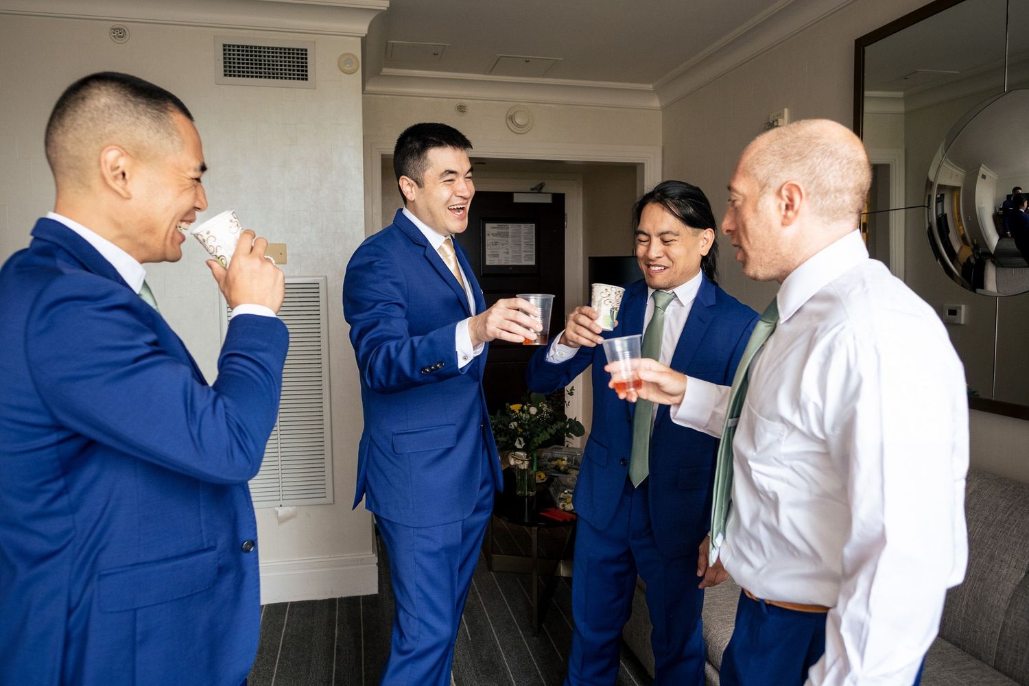 Four groomsmen in blue suits toasting with small cups in hotel room before wedding ceremony.
