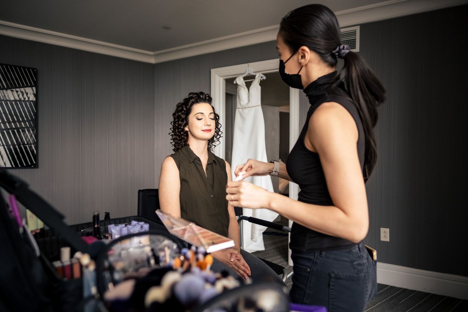 Makeup artist in black applying products to bride with curly hair before wedding ceremony.
