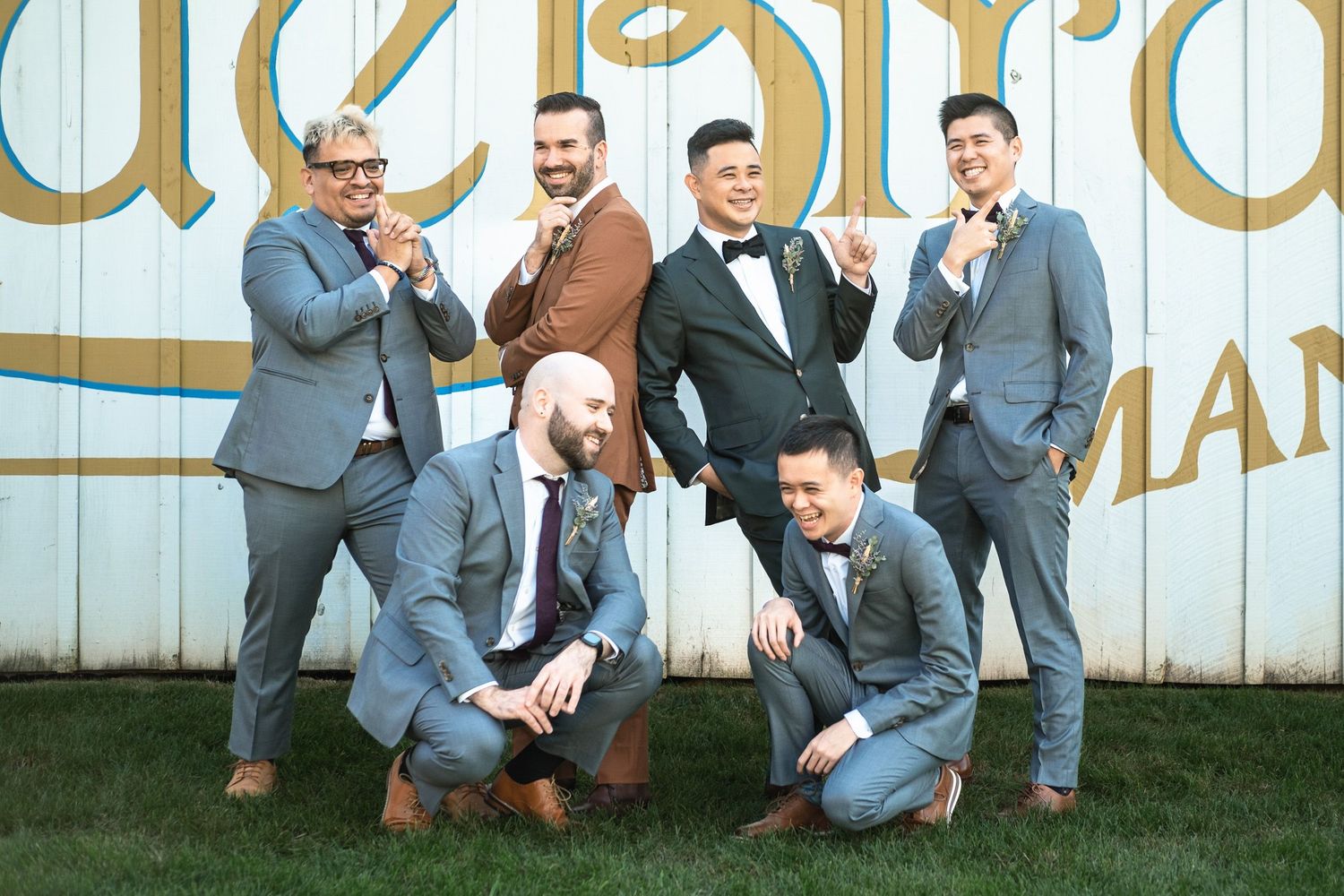 Six groomsmen pose playfully in front of Blue Bird Manor barn sign at wedding.
