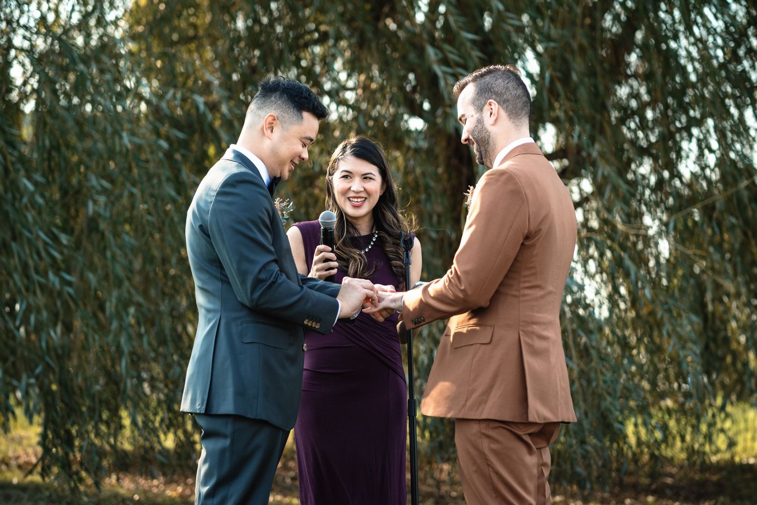 Two grooms exchange rings during an outdoor wedding ceremony officiated by a woman in a purple dress.