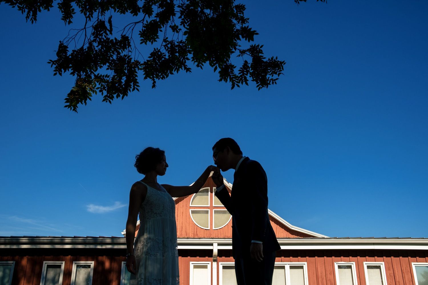 Romantic silhouette of bride and groom in front of a red barn under a deep blue sky at golden hour.