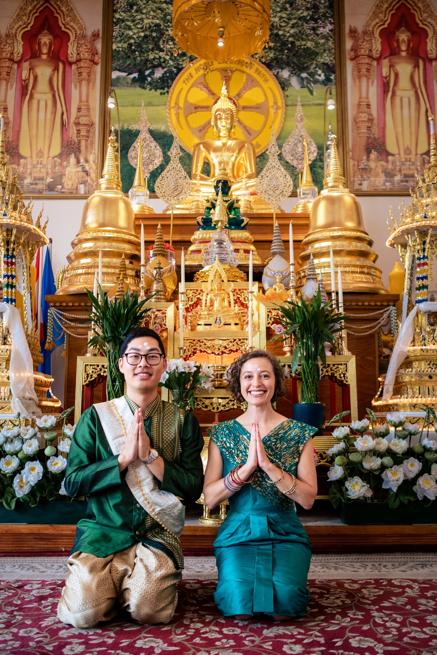 Couple in traditional Thai green silk attire performs wai gesture before golden Buddha altar in temple.