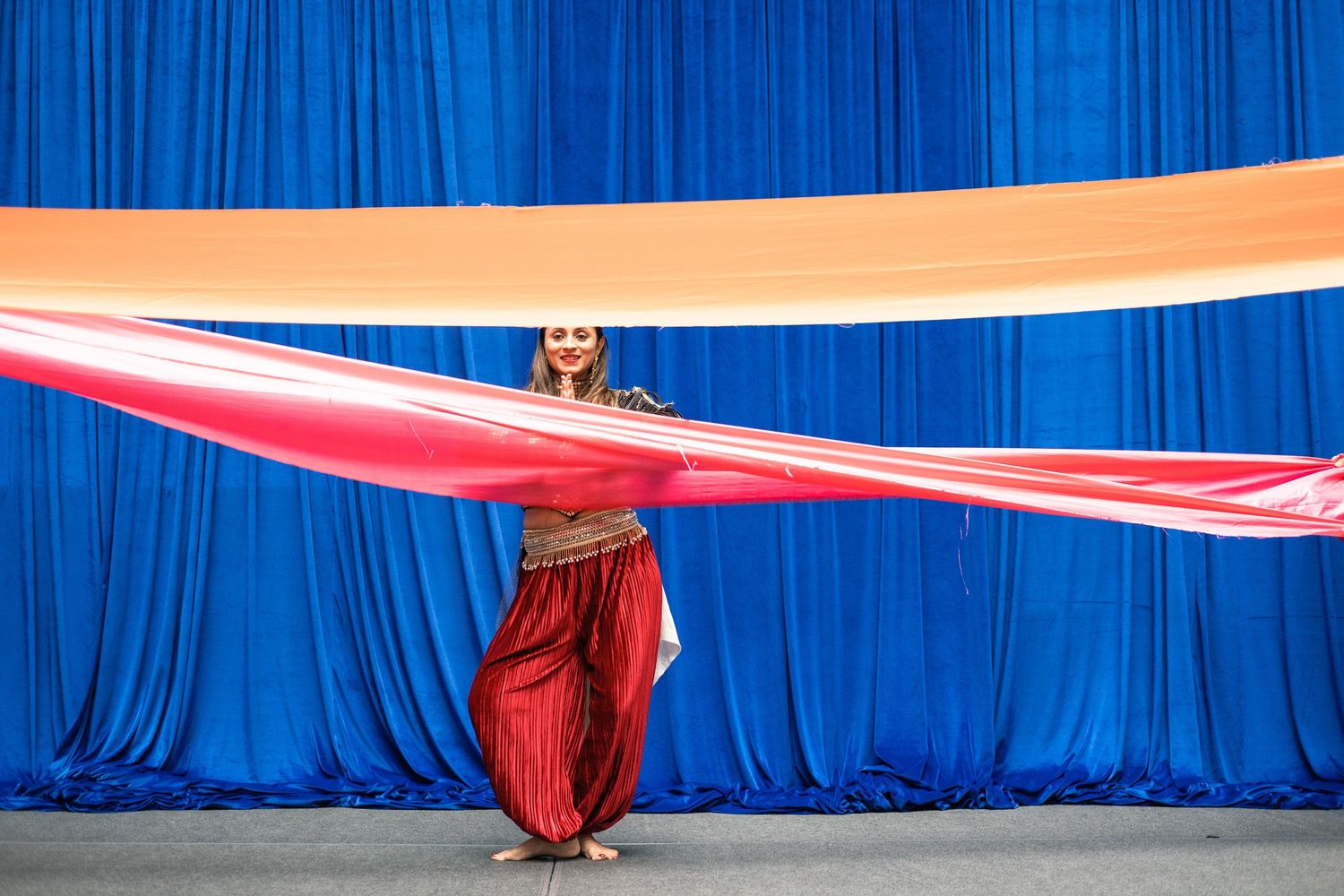A belly dancer in red harem pants holds a flowing orange and red silk veil against a blue curtain backdrop.